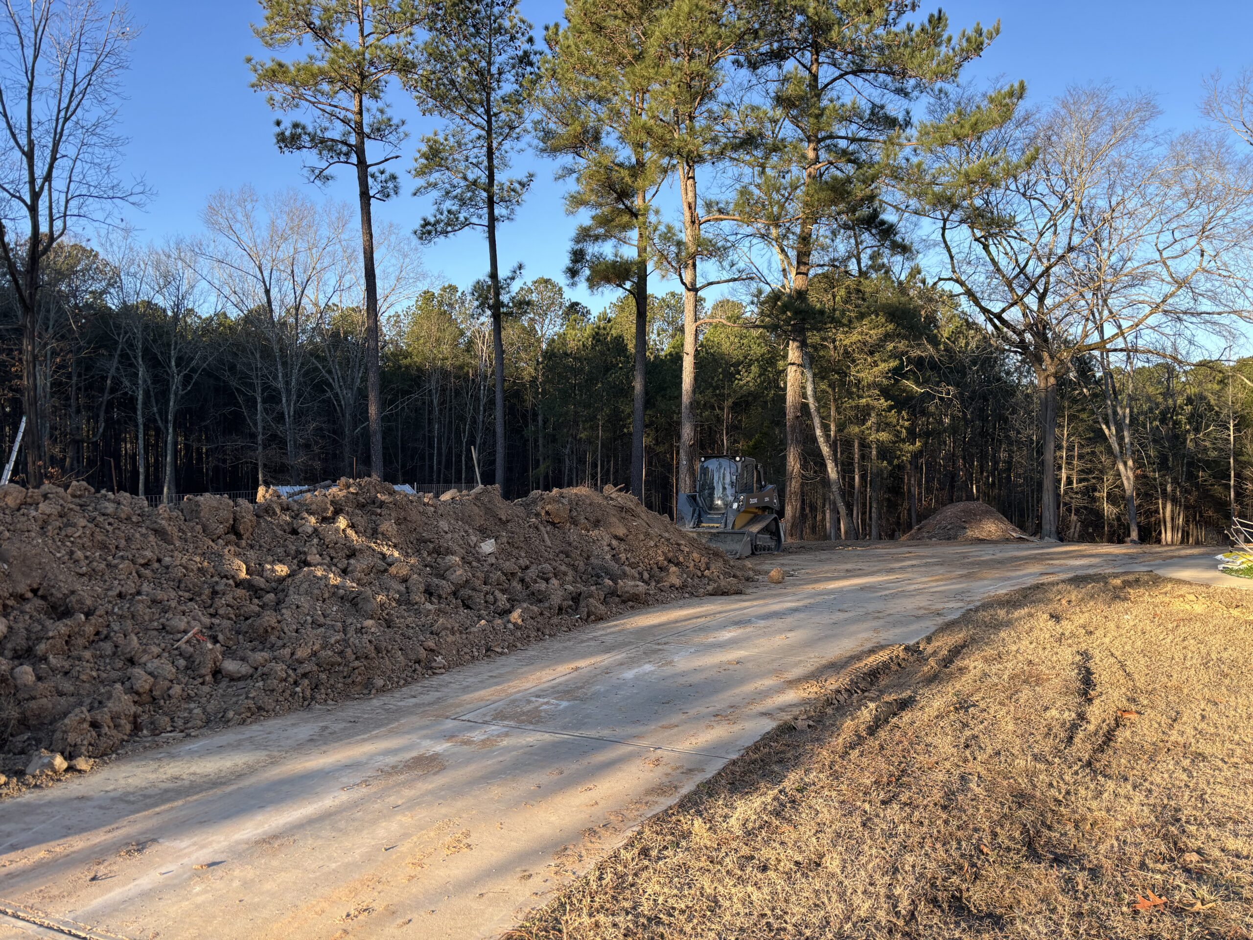 A dirt pile and excavator sit next to a paved driveway near a wooded area with tall pine trees on a clear day.