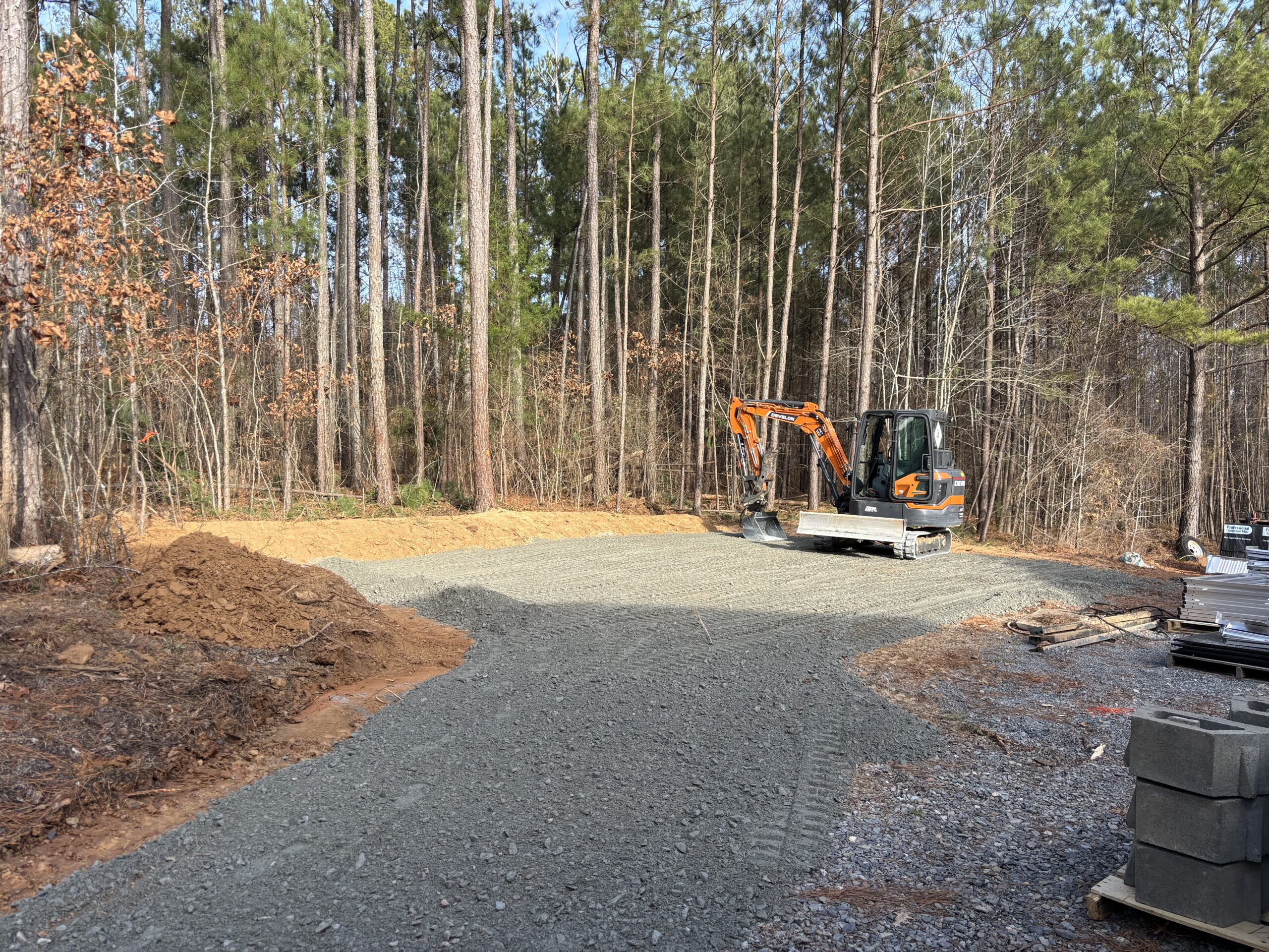 A small excavator is parked on a freshly laid gravel surface in a clearing surrounded by trees, with construction materials stacked nearby.