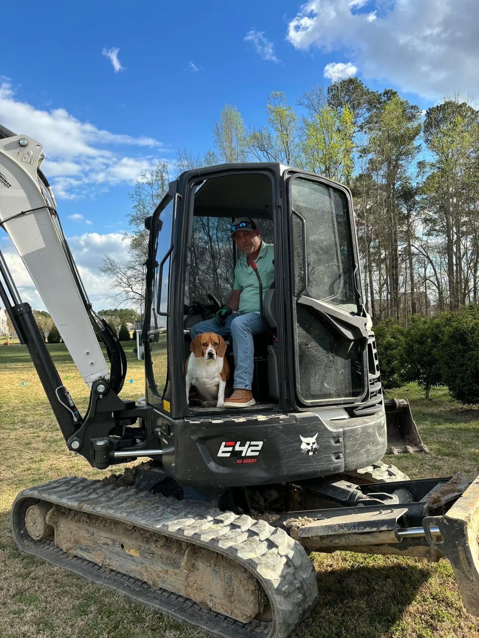 A man and a dog sit together inside the cab of a small excavator parked on grass, with trees and a partly cloudy sky in the background.