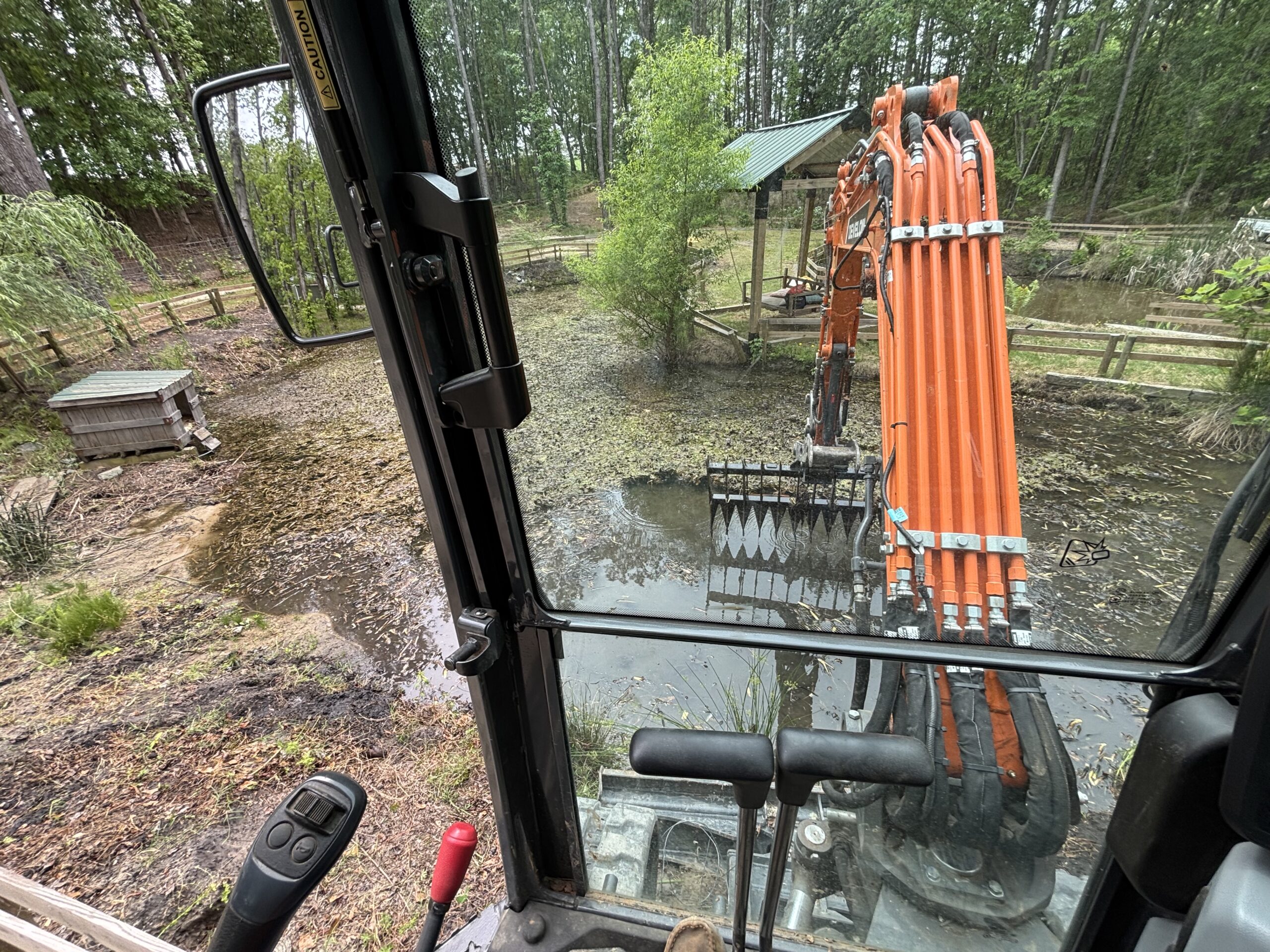 View from inside an excavator cab showing the machine's orange arm reaching toward a muddy, partially flooded area with a shed and fence in the background.