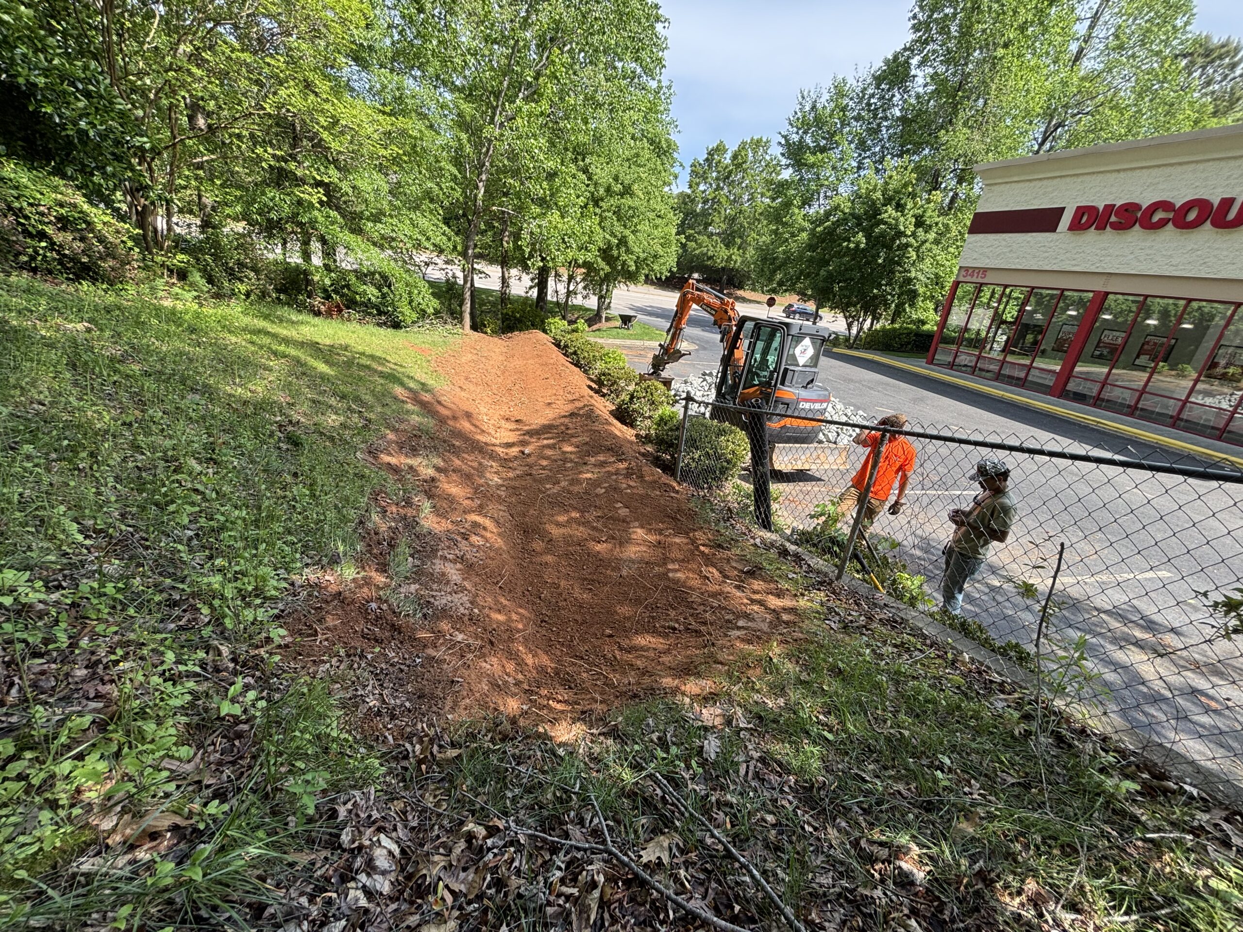 A recently cleared dirt path slopes down toward a chain-link fence near a parking lot, with construction workers and equipment visible by a discount store.