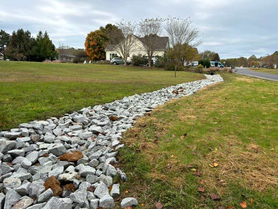 A line of gray rocks forms a drainage ditch beside a grassy area near a suburban house and a road on an overcast day.