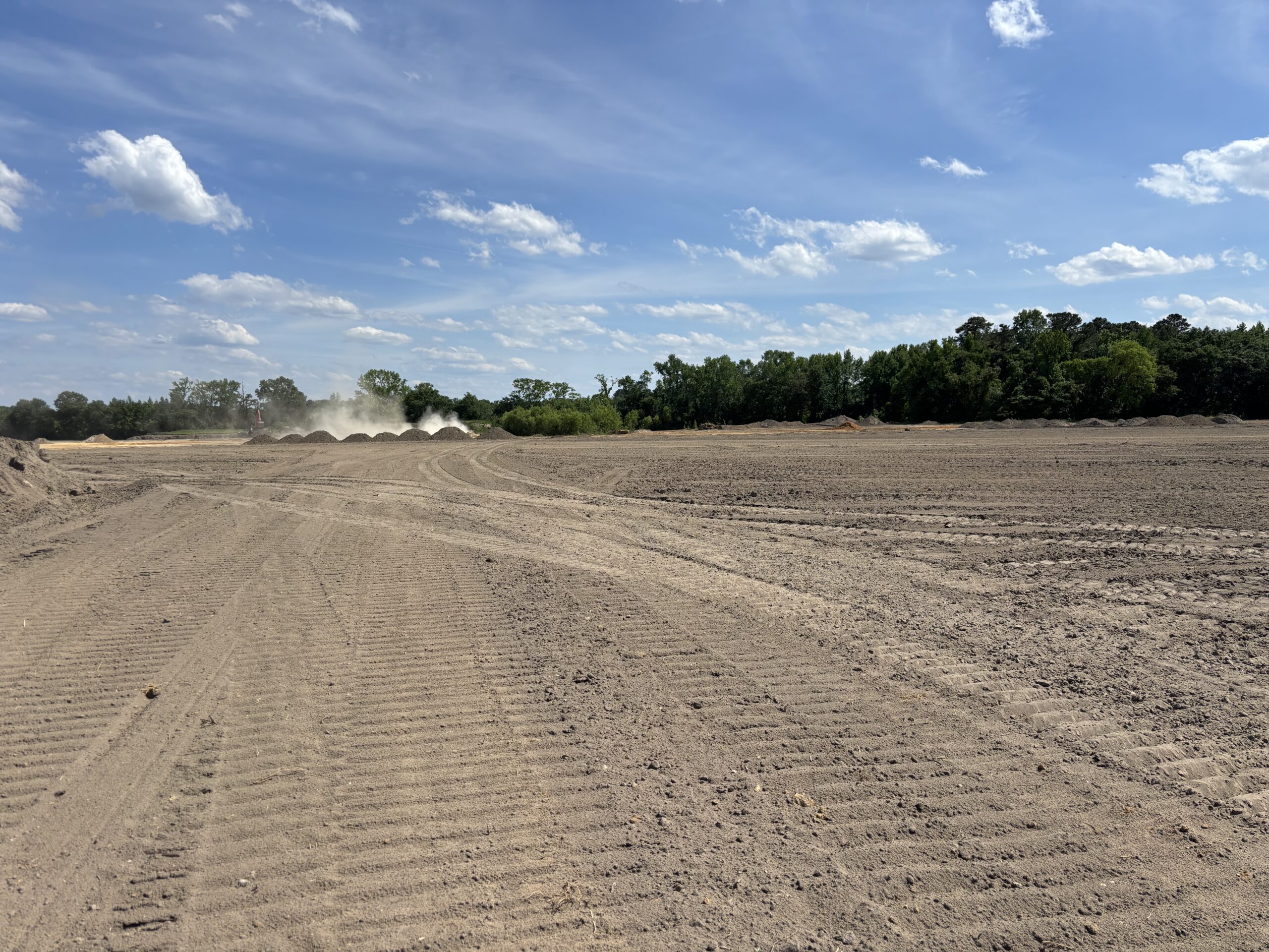 Expansive, freshly-leveled dirt field with tire tracks and a distant tree line under a partly cloudy sky; dust clouds are visible near the horizon.
