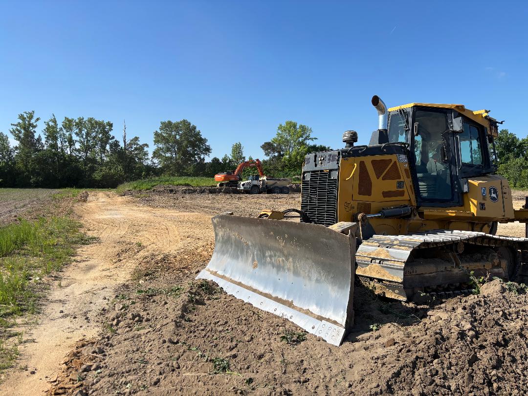 A yellow bulldozer sits on a dirt construction site, ready for grading, with an excavator and more equipment in the background under a clear blue sky.
