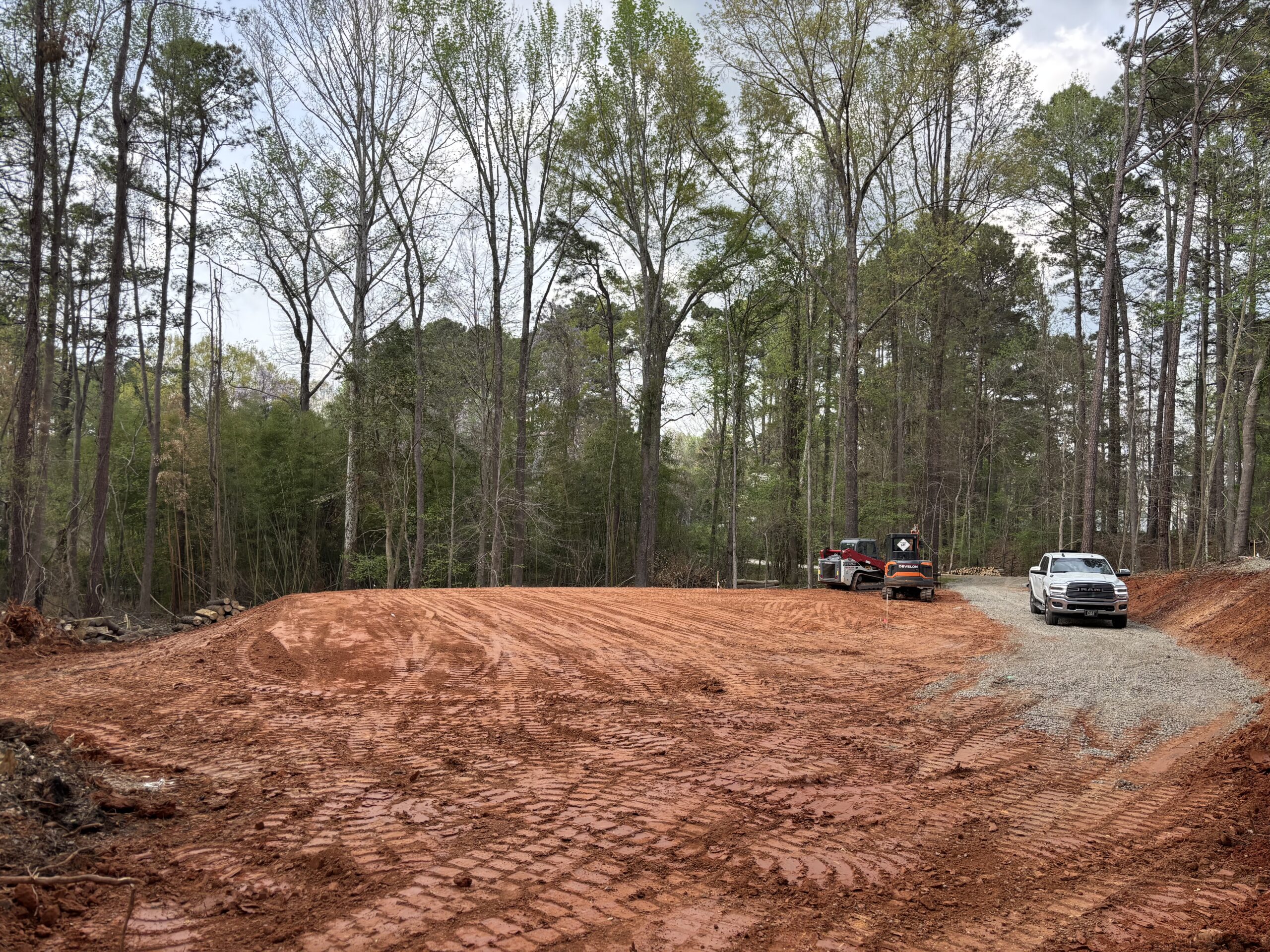 A freshly cleared and leveled red dirt area in a forest, with two pickup trucks parked on a gravel section to the right under tall trees.