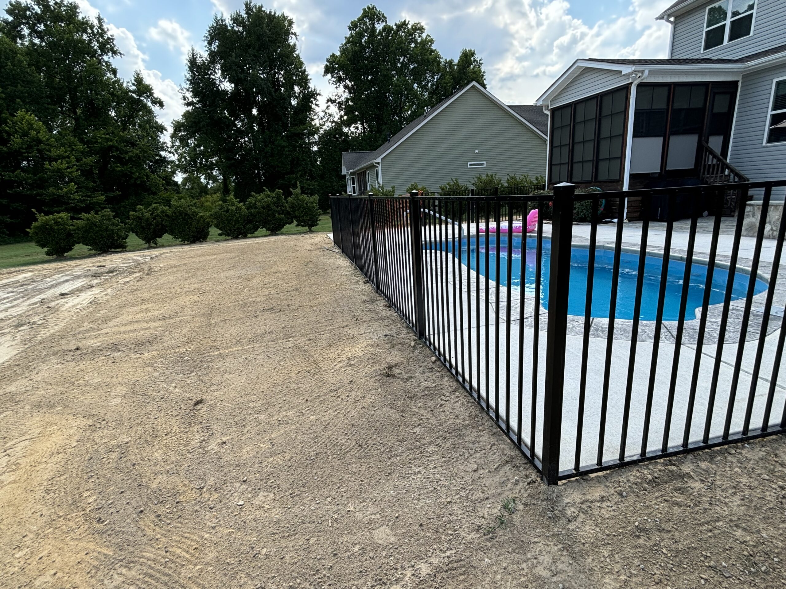 A fenced-in backyard pool next to a house with bare dirt and some grass on the surrounding yard; trees are visible in the background.