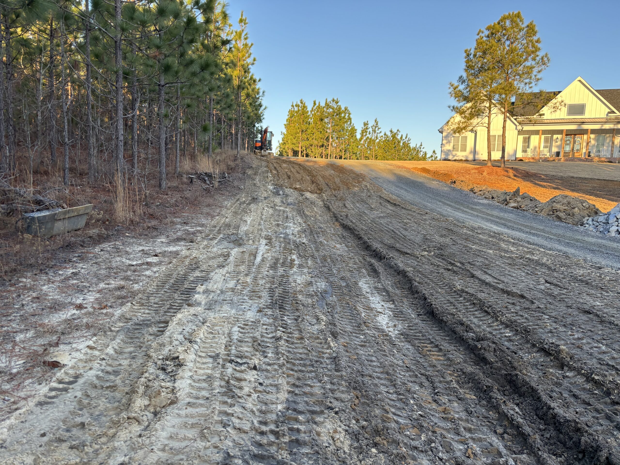 A dirt and gravel road with visible tire tracks leads uphill toward a house, bordered by pine trees under a clear sky.