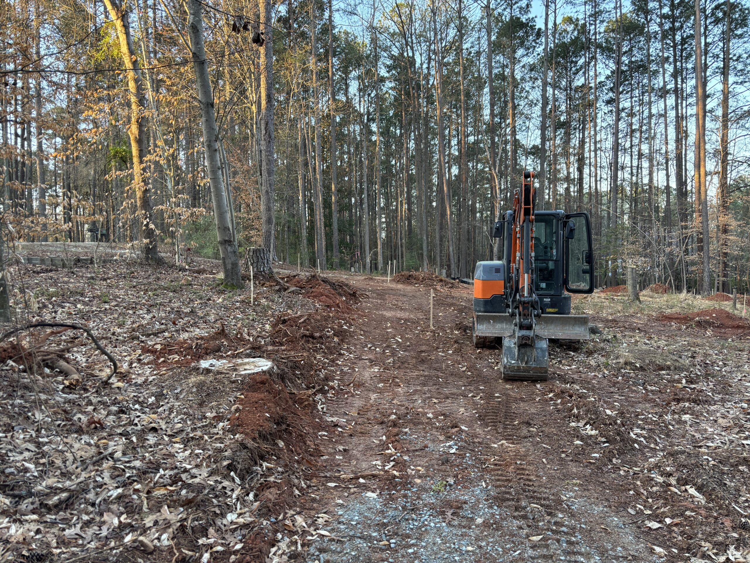 Excavator parked on a cleared dirt path in a forested area with trees and scattered leaves surrounding the construction site.