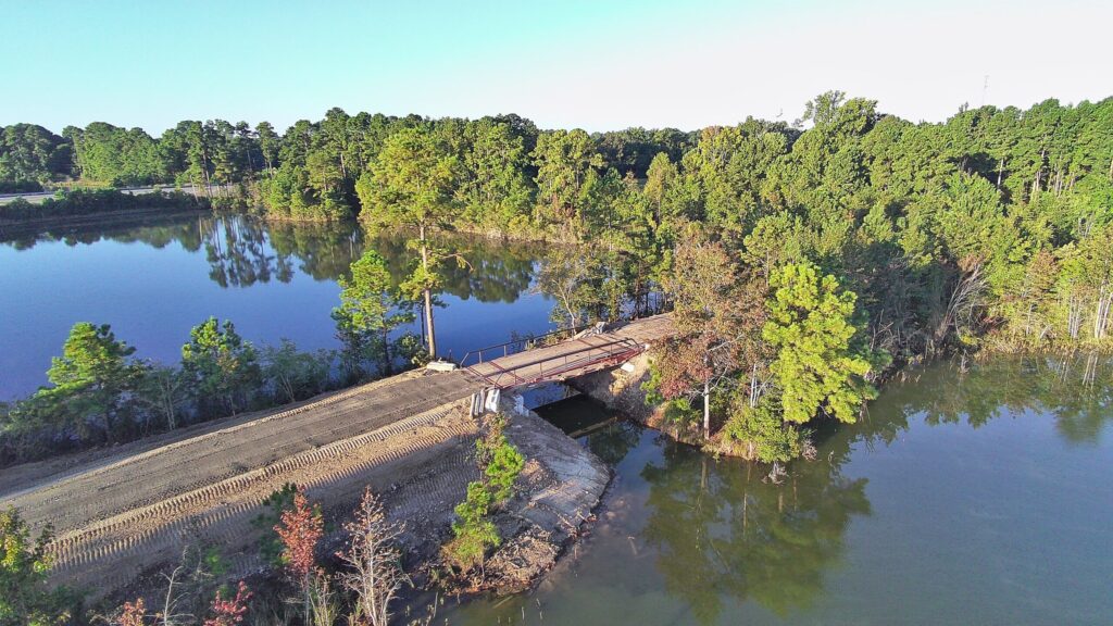 A dirt road with recent grading and a small bridge crosses over water, surrounded by dense green trees and reflecting waters under a clear sky.