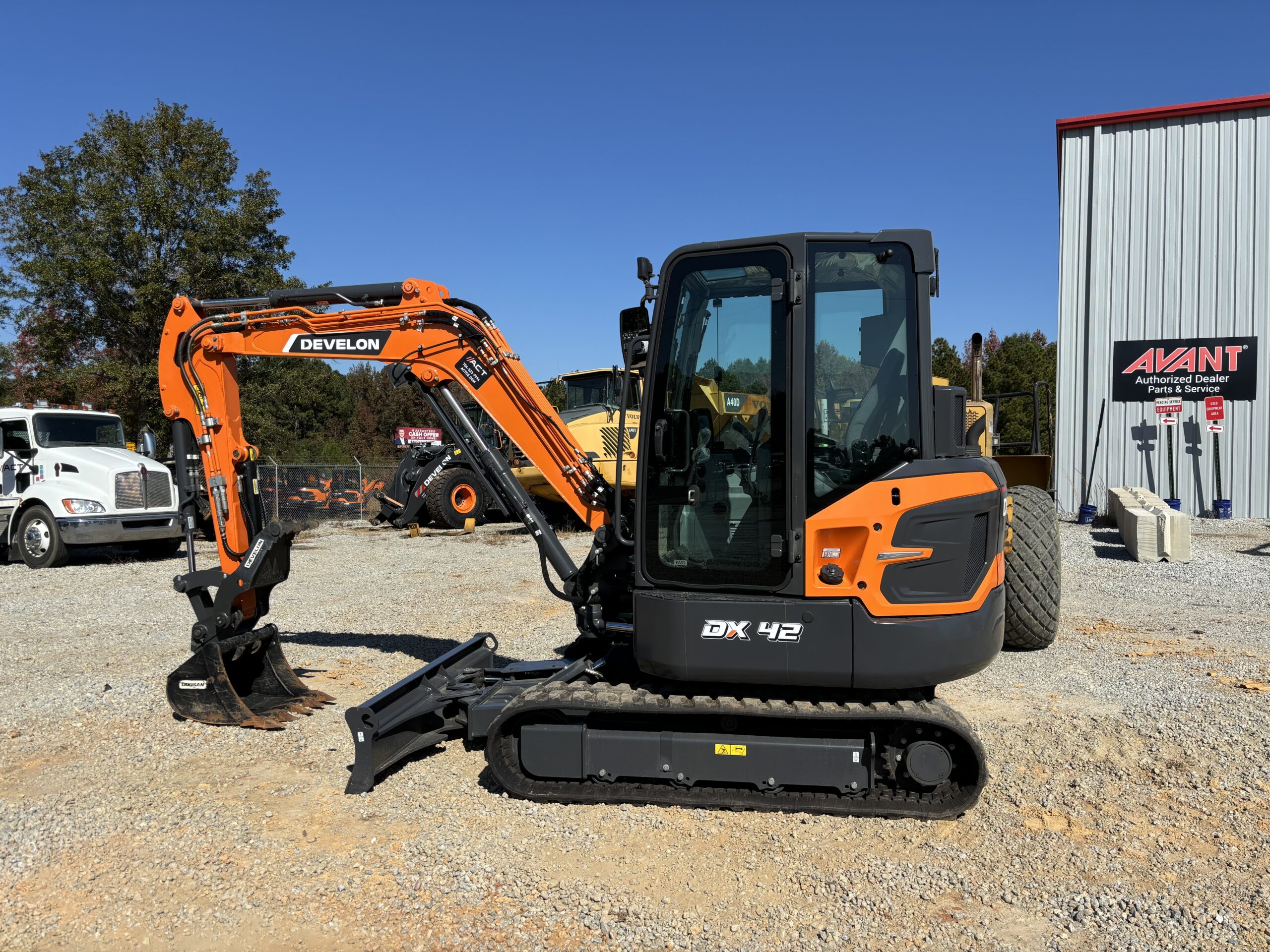 An orange Develon DX42 compact excavator, available for excavator rental, is parked on gravel near a metal building and other construction vehicles under a clear blue sky.