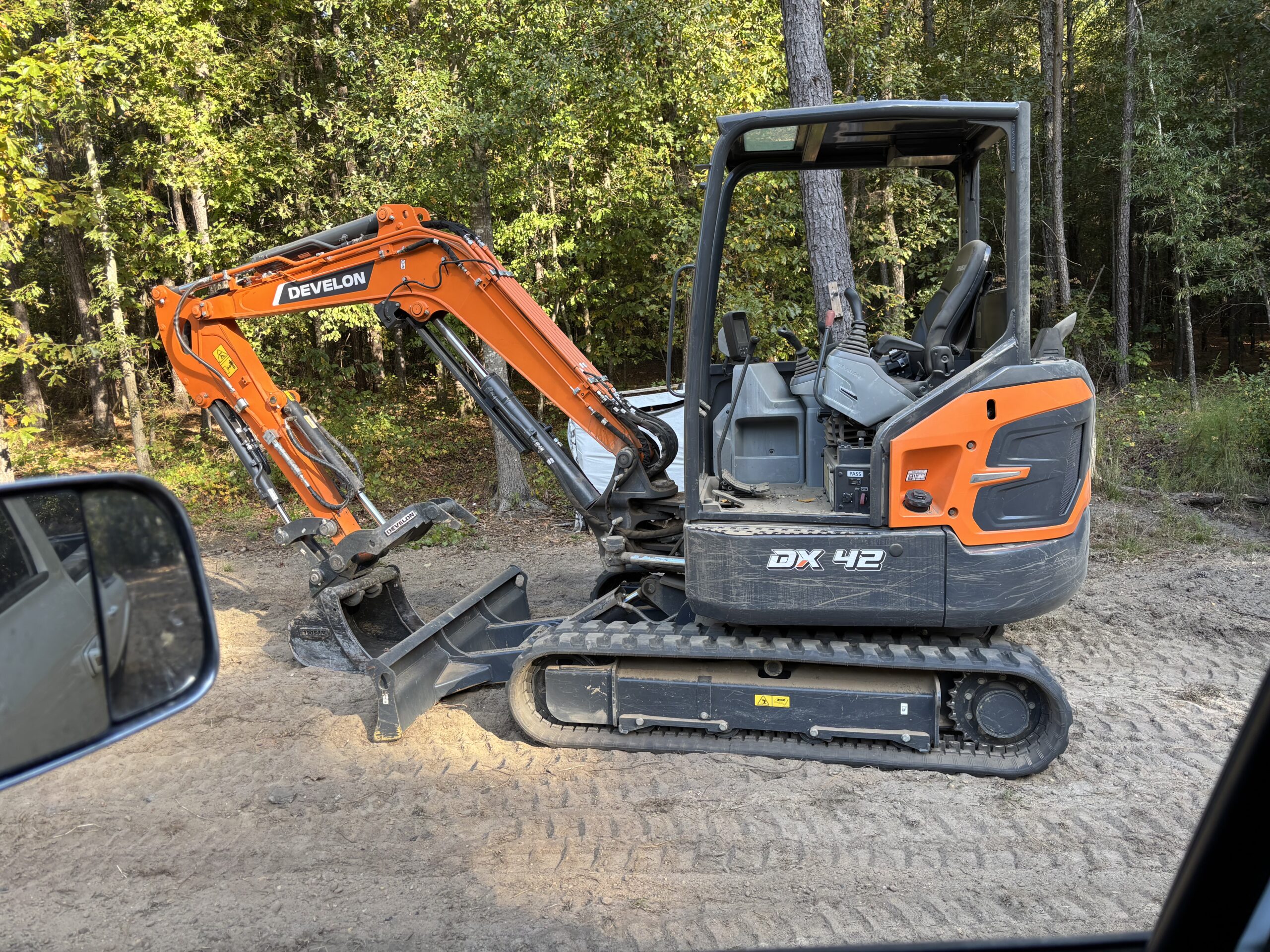 An orange Develon DX42 mini excavator with a bucket attachment, possibly available for excavator rental, is parked on a dirt surface near trees, viewed from a nearby vehicle.